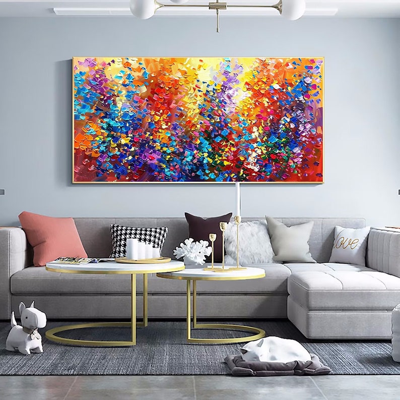 May include: A large abstract painting with a colorful, textured design. The painting features a variety of colors, including blue, green, yellow, orange, and red. The painting is framed in a gold frame and is hanging on a wall in a living room.