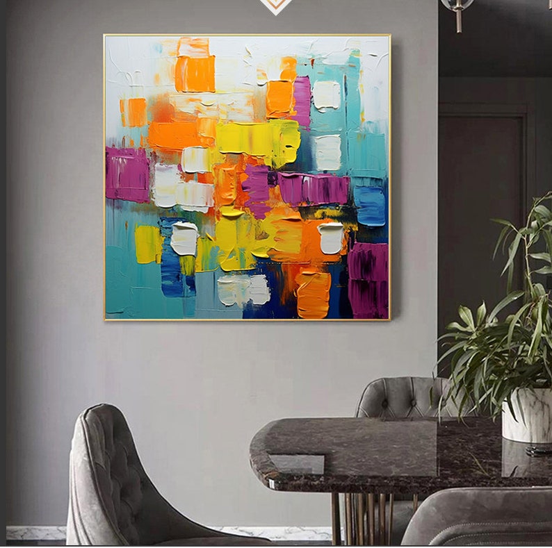 May include: Abstract painting with a colorful geometric design. The painting features squares and rectangles in shades of yellow, orange, blue, purple, and white. The painting is framed in gold.