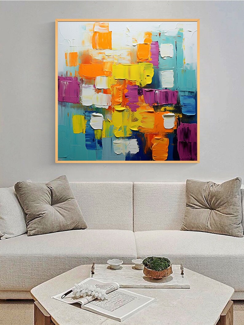 May include: Abstract painting with a colorful geometric design. The painting features squares and rectangles in shades of blue, yellow, orange, purple, and white. The painting is framed in a wooden frame.
