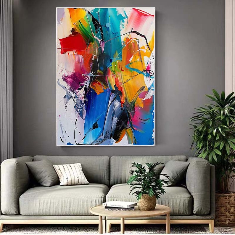 Puede incluir: Pintura abstracta con trazos audaces de azul, amarillo, rojo, rosa y verde sobre un fondo blanco. La pintura est&aacute; enmarcada y colgada en una pared gris sobre un sof&aacute; gris.