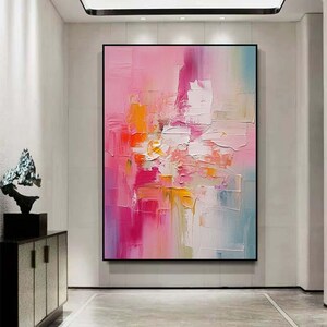 K&ouml;nnte beinhalten: Abstraktes &Ouml;lgem&auml;lde mit schwarzem Rahmen. Das Kunstwerk zeigt eine lebendige Mischung aus Farben, darunter Rosa, Orange, Wei&szlig; und Blau. Die Pinselstriche sind dick und strukturiert und schaffen ein dynamisches und ausdrucksstarkes Kunstwerk. Das Gem&auml;lde wird in einem modernen Innenraum ausgestellt.