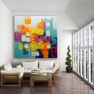 May include: Abstract painting with a colorful geometric design. The painting features squares and rectangles in shades of yellow, orange, blue, purple, and white. The painting is framed and hanging on a white wall.
