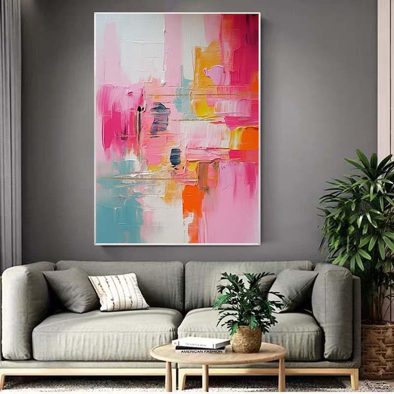K&ouml;nnte beinhalten: Abstraktes Gem&auml;lde mit wei&szlig;em Rahmen. Das Kunstwerk zeigt kr&auml;ftige Pinselstriche in Rosa-, Orange-, Gelb- und Blaut&ouml;nen. Das Gem&auml;lde wird &uuml;ber einem grauen Sofa mit Kissen, einem Holztisch und einer Topfpflanze ausgestellt.