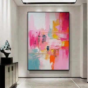 K&ouml;nnte beinhalten: Abstrakte Kunstmalerei mit schwarzem Rahmen. Das Kunstwerk zeigt eine lebendige Mischung aus Farben, darunter Rosa, Orange, Gelb und Blau, die mit dicken Pinselstrichen aufgetragen wurden. Das Gem&auml;lde wird in einem hellen Flur ausgestellt.