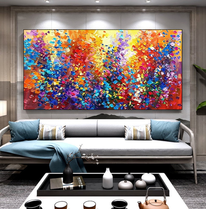 May include: Abstract painting with a colorful, textured, and vibrant design. The painting features a mix of blue, yellow, red, purple, and green colors. The painting is displayed on a wall in a living room setting.