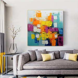 May include: Abstract painting with a colorful geometric design. The painting features squares and rectangles in shades of yellow, orange, blue, purple, and white. The painting is framed in a gold frame.