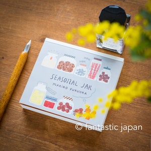 Op de afbeelding: Een grijs en wit geïllustreerd notitieboekje met de tekst "Seasonal Jar Mariko Fukuoka" en een tekening van potten gevuld met verschillende items zoals koekjes, snoep en fruit. Het notitieboekje ligt op een houten oppervlak met een gele bloem en een zwarte inktpot.