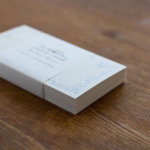 May include: A stack of blank white note cards with a blue floral design on the edge. The cards are wrapped in a white paper band with the text "The Little Note Card Company" printed on it.