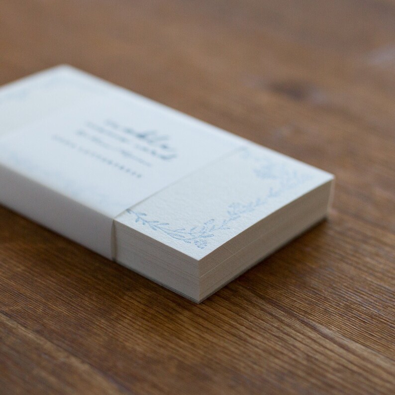 May include: A stack of blank white note cards with a blue floral design on the edge. The cards are wrapped in a white paper band with the text "The Little Note Card Company" printed on it.