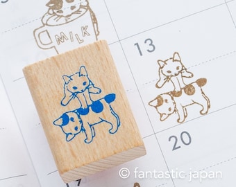 May include: Wooden stamp with a blue ink design of two cats stacked on top of each other. The top cat is sitting on the bottom cat's back. The bottom cat is walking. The stamp is on a white calendar page with the number 13 written on it.