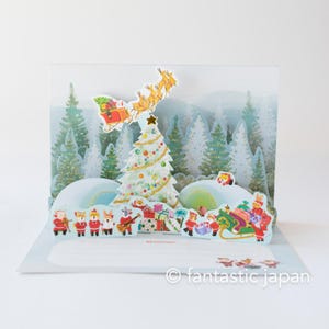May include: A pop-up Christmas card featuring a decorated Christmas tree, Santa's sleigh, and various festive characters. The background depicts a snowy forest scene. The card is white and blue with colorful accents.
