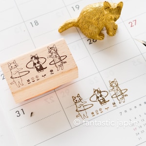 Może przedstawiać: Drewniana pieczątka z trzema kotami robiącymi hula-hoop, odbita na kalendarzu. W pobliżu znajduje się mała złota figurka kota. Projekt pieczątki zawiera kwiaty i tekst "fantastic japan".