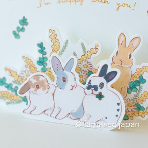 May include: A pop-up card featuring four adorable bunnies surrounded by yellow and green flowers. The card has the text "I'm happy with you!" written on it.