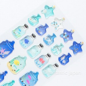 Puede incluir: Una hoja de pegatinas azules y blancas con varios dise&ntilde;os de tem&aacute;tica marina. Las pegatinas tienen forma de botellas y estrellas, y cada botella contiene una criatura marina diferente, como un pez, una medusa o una estrella de mar. Las pegatinas son perfectas para a&ntilde;adir un toque de fantas&iacute;a a tus manualidades, diarios o planificadores.