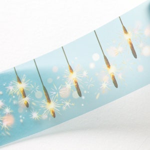 May include: A roll of washi tape featuring a repeating pattern of gold sparklers against a light blue background. The tape has a diagonal orientation, with the sparklers appearing to be lit. The design includes white bokeh effects.