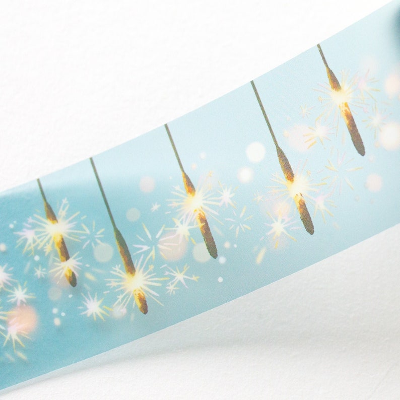 May include: A roll of washi tape featuring a repeating pattern of gold sparklers against a light blue background. The tape has a diagonal orientation, with the sparklers appearing to be lit. The design includes white bokeh effects.