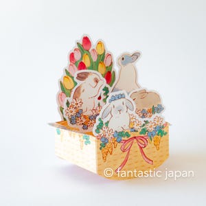 May include: A pop-up card featuring a yellow basket with a pink ribbon, filled with colorful tulips, daisies, and three white rabbits. The card is decorated with a green vine pattern and the text "fantastic japan" is printed on the bottom.