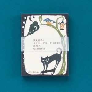 May include: A rectangular card with a black cat illustration, a bird, and a lantern on a tree branch. The card has Japanese text and is held together with a clear band. The background is a dark blue.