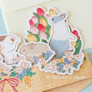 May include: A pop-up card featuring three bunnies and a bouquet of tulips. The bunnies are white with gray, brown, and pink accents. The tulips are pink, yellow, and red. The card is decorated with white and blue flowers. The text "&copy; fantastic japan" is printed on the bottom of the card.