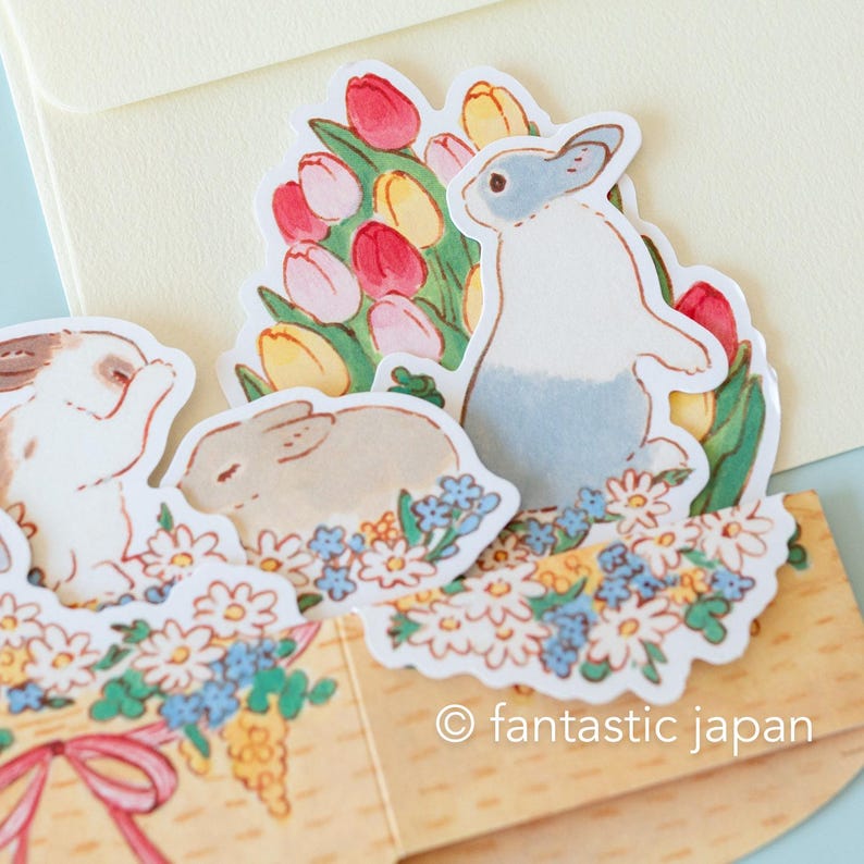 May include: A pop-up card featuring three bunnies and a bouquet of tulips. The bunnies are white with gray, brown, and pink accents. The tulips are pink, yellow, and red. The card is decorated with white and blue flowers. The text "&copy; fantastic japan" is printed on the bottom of the card.