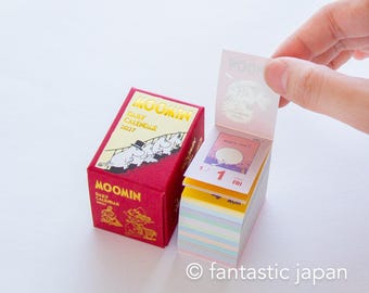 Peut inclure: Un calendrier quotidien Moomin rouge pour 2027 avec des lettres dorées et des illustrations de personnages sur la boîte. À côté, une pile de feuilles de calendrier détachables indique la date du vendredi 1er janvier avec un imprimé coloré.