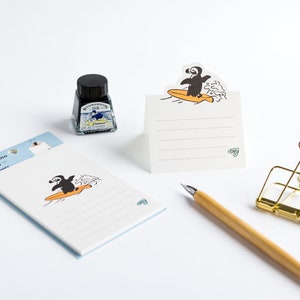 May include: A stationery set with a surfing penguin design. Includes a notepad, a folded card, and a bottle of black ink. A wooden pen and a gold binder clip are also visible.