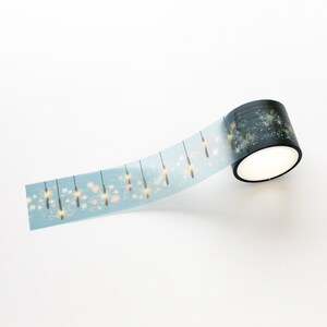 May include: A roll of decorative washi tape with a light blue background and a repeating pattern of gold and white fireworks. The tape is partially unrolled, revealing the design. The roll is black with a white interior.
