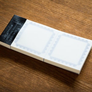 Puede incluir: Un bloc de notas blanco con dos p&aacute;ginas en blanco, cada una con un borde decorativo azul. El bloc de notas tiene una etiqueta negra en la parte delantera que dice "LABEL SAMPLE PRINTING PROOF #203119-02".