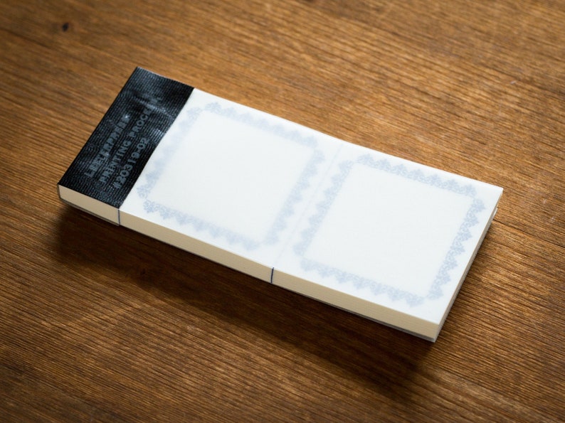 Puede incluir: Un bloc de notas blanco con dos p&aacute;ginas en blanco, cada una con un borde decorativo azul. El bloc de notas tiene una etiqueta negra en la parte delantera que dice "LABEL SAMPLE PRINTING PROOF #203119-02".