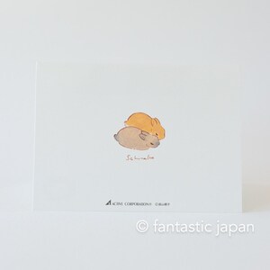 May include: A postcard featuring a watercolour illustration of two small rabbits sleeping on top of each other. The rabbits are brown and orange. The text "Schnuggle" is written in red below the rabbits.