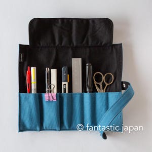 Puede incluir: Un estuche enrollable de lona azul con solapa negra, que contiene varios artículos de papelería. Incluye bolígrafos rojos y amarillos, un bolígrafo negro, una goma de borrar blanca, un clip rosa, un rotulador azul y naranja, una regla metálica, un bolígrafo negro y tijeras doradas. El texto "© fantastic japan" es visible.