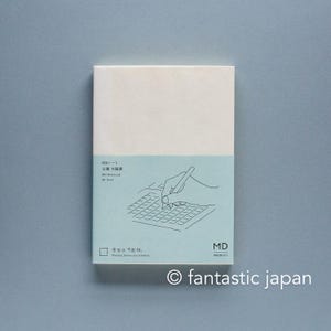 May include: A rectangular notebook with a cream-colored cover and a light blue paper band. The band features a line drawing of a hand writing on a grid, with text including MD Notebook, A6 Grid, and the phrase Pacing moves you forward.