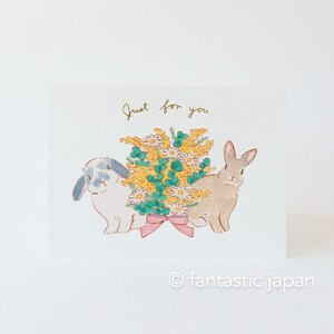 May include: A greeting card featuring a watercolour illustration of two rabbits holding a bouquet of yellow and white flowers. The card has the text "Just for you" written in gold lettering.