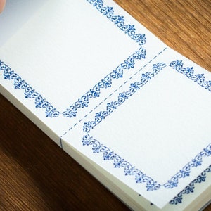 Puede incluir: Un bloc de notas blanco con bordes decorativos azules. El bloc de notas est&aacute; doblado por la mitad y tiene dos p&aacute;ginas en blanco.
