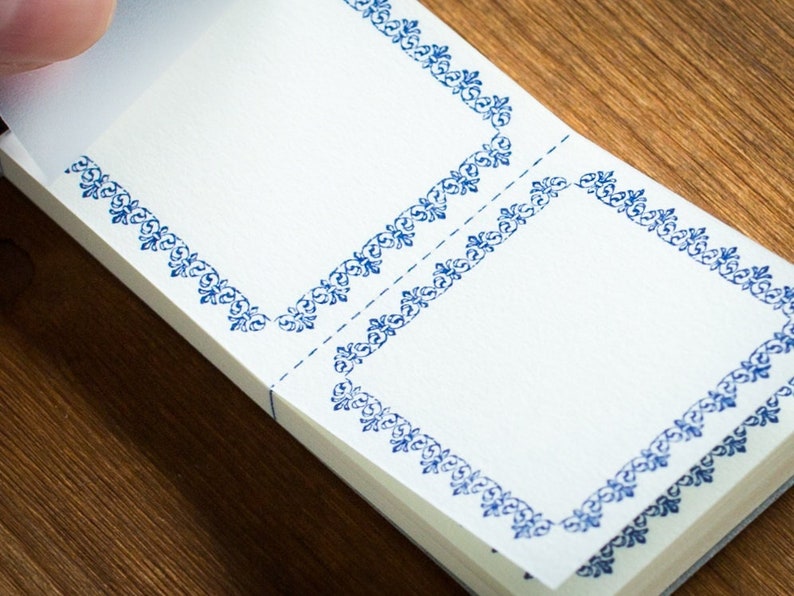 Puede incluir: Un bloc de notas blanco con bordes decorativos azules. El bloc de notas est&aacute; doblado por la mitad y tiene dos p&aacute;ginas en blanco.