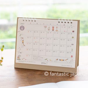 May include: A 2027 desk calendar with a spiral binding and a brown cardboard stand. The page displays the month of January with a grid layout, Japanese text, and illustrations of cats playing with toys, flowers, and traditional New Year items.