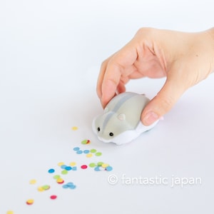 Pode incluir: Um pequeno brinquedo de hamster cinza e branco com olhos pretos e uma faixa cinza nas costas. O brinquedo está sendo segurado por uma mão e está rodeado de confete colorido.