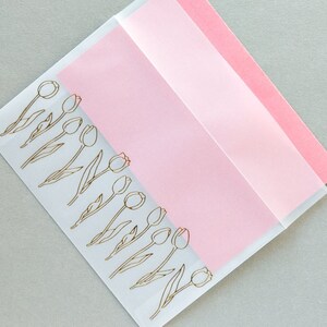 Puede incluir: Un sobre transparente con un inserto de papel rosa. El sobre tiene un contorno dorado de una fila de tulipes.