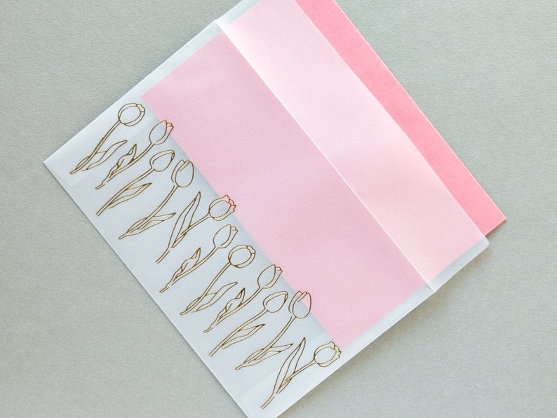 Puede incluir: Un sobre transparente con un inserto de papel rosa. El sobre tiene un contorno dorado de una fila de tulipes.