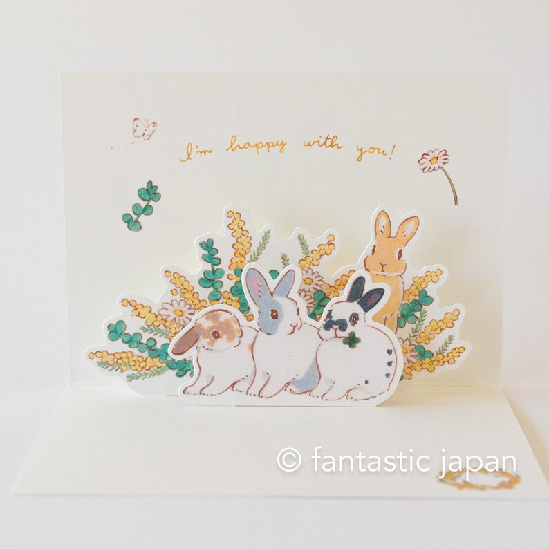May include: A pop-up card featuring a 3D scene of four bunnies surrounded by yellow flowers and green leaves. The card reads "I'm happy with you!" and is signed "fantastic japan".