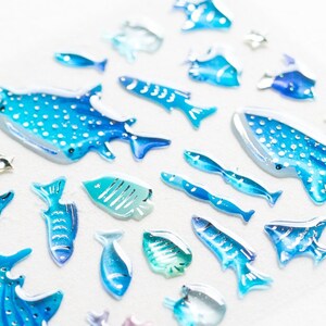 May include: A collection of blue and white whale shark and fish stickers. The stickers are made of a clear, glossy material and have a 3D effect.