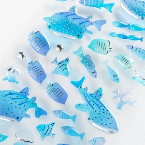 May include: A collection of blue and silver fish-shaped stickers. The stickers are arranged in a diagonal line on a white background. The fish are all different sizes and shapes, and some have glitter accents.