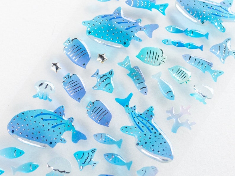 May include: A collection of blue and silver fish-shaped stickers. The stickers are arranged in a diagonal line on a white background. The fish are all different sizes and shapes, and some have glitter accents.