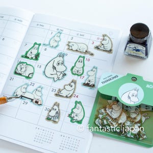 Puede incluir: Pegatinas de personajes de Mumin en varias poses, dispuestas en un calendario. Las pegatinas son blancas con contornos negros y bordes verdes. También se ve un frasco de tinta azul y un paquete de pegatinas con "FACE SERIES THINKING".