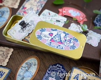 May include: A gold metal tin filled with colorful stickers featuring Alice in Wonderland characters and designs. The stickers are in a variety of shapes and sizes, including oval, rectangular, and square. The tin is open and the stickers are visible inside. The text "Heart" and "Shinzi Katoh" are visible on the stickers.