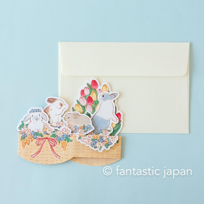 May include: A pop-up card featuring a basket of flowers with four bunnies. The bunnies are white, gray, and brown. The flowers are pink, yellow, and white. The card is made of paper and has a white envelope.