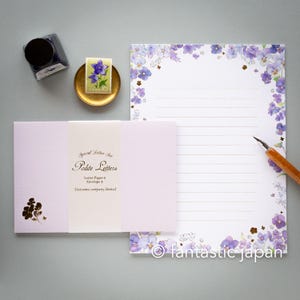 Może przedstawiać: Zestaw papierniczy z lawendową kopertą i liniowanym papierem listowym, oba zdobione delikatnymi fioletowymi wzorami kwiatowymi. Znaczek z fioletowym kwiatem znajduje się na złotej miseczce, obok atramentu i pióra.