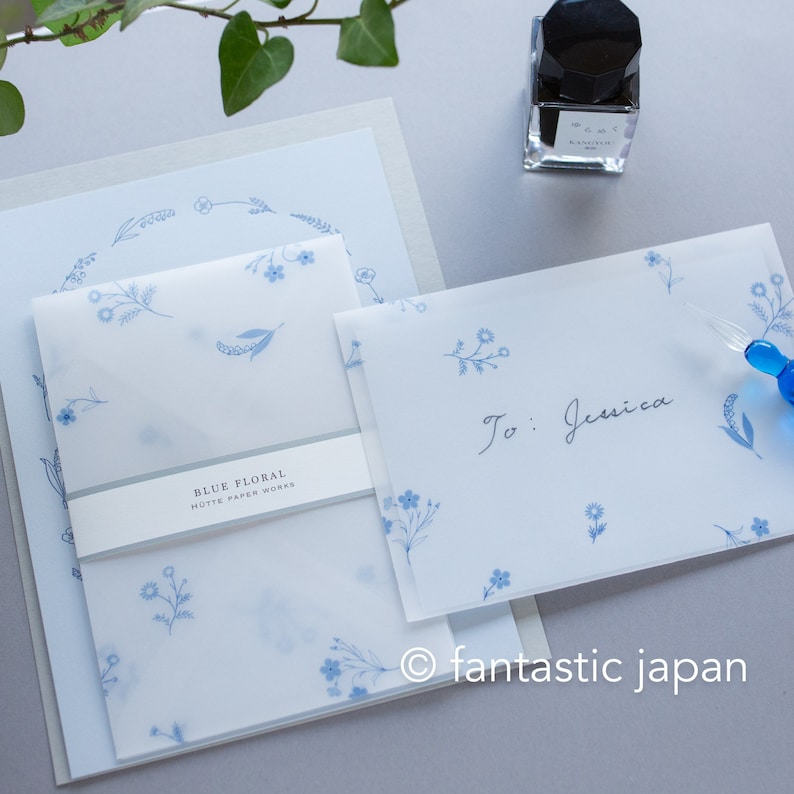 Pode incluir: Conjunto de papelaria com desenhos florais azuis. O conjunto inclui envelopes, cart&otilde;es e um envelope transl&uacute;cido com o texto "BLUE FLORAL HUTTE PAPER WORKS". Um frasco de tinta preta e uma caneta de vidro tamb&eacute;m s&atilde;o vis&iacute;veis.