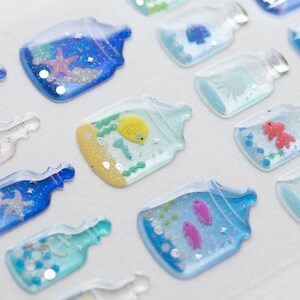 Puede incluir: Surtido de pegatinas de botellas de vidrio transparente con purpurina y peces de colores en su interior. Las pegatinas son 3D y tienen un acabado brillante.