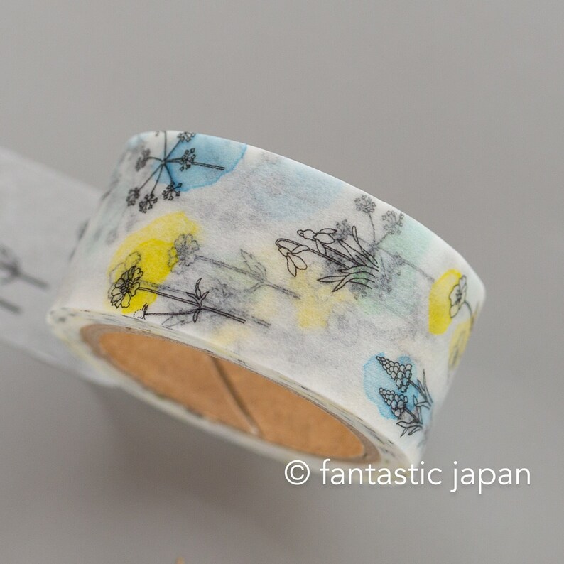 Puede incluir: Un rollo de cinta washi blanca con un dise&ntilde;o floral. La cinta presenta contornos negros de flores y hojas con acentos azul y amarillo como de acuarela. La cinta est&aacute; en un carrete de cart&oacute;n marr&oacute;n.